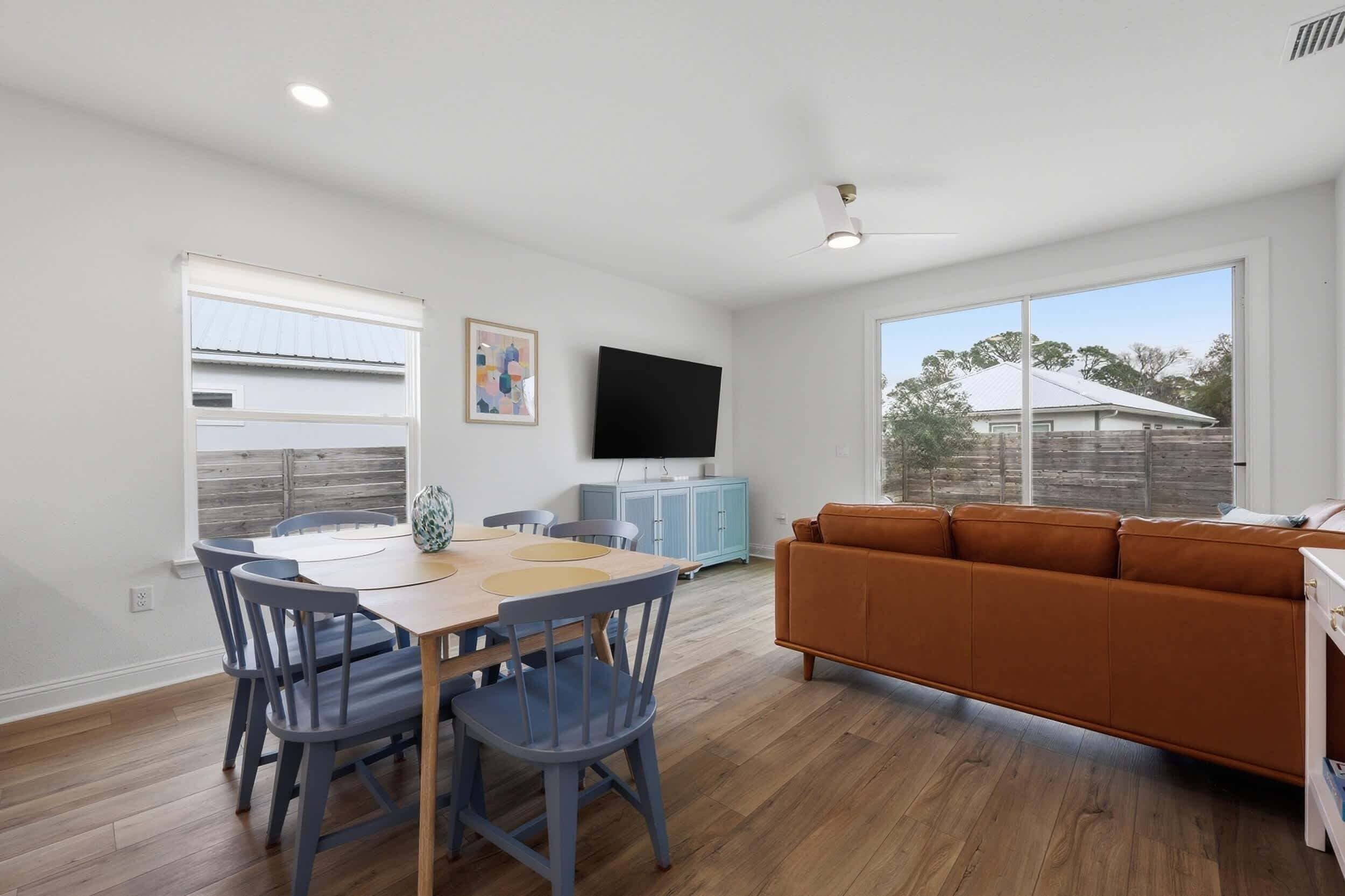 A modern living room with a wooden dining table, blue chairs, a brown sofa, a flat-screen TV, and a large window.