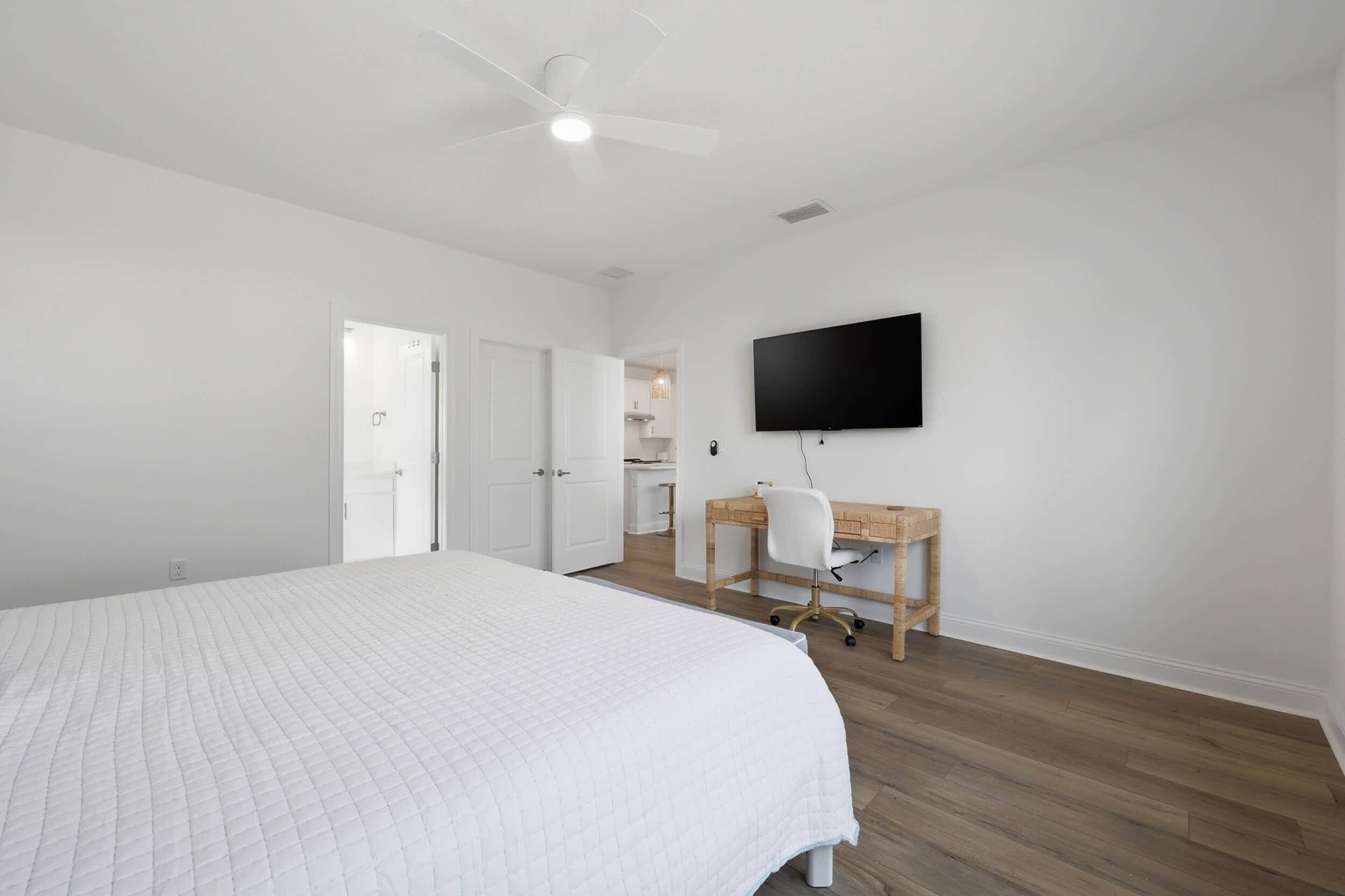 A bedroom with a bed, a desk, a chair, a ceiling fan, and a TV on the wall.