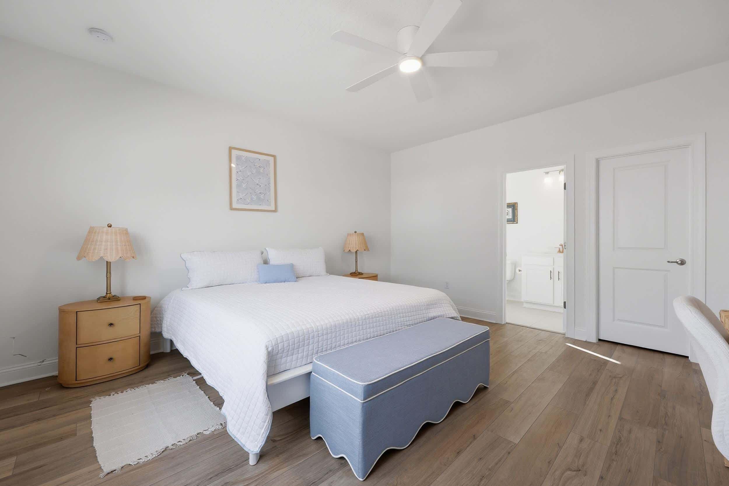 A bedroom with a bed, bedside tables, a nightstand, a bench, a ceiling fan, and a door.