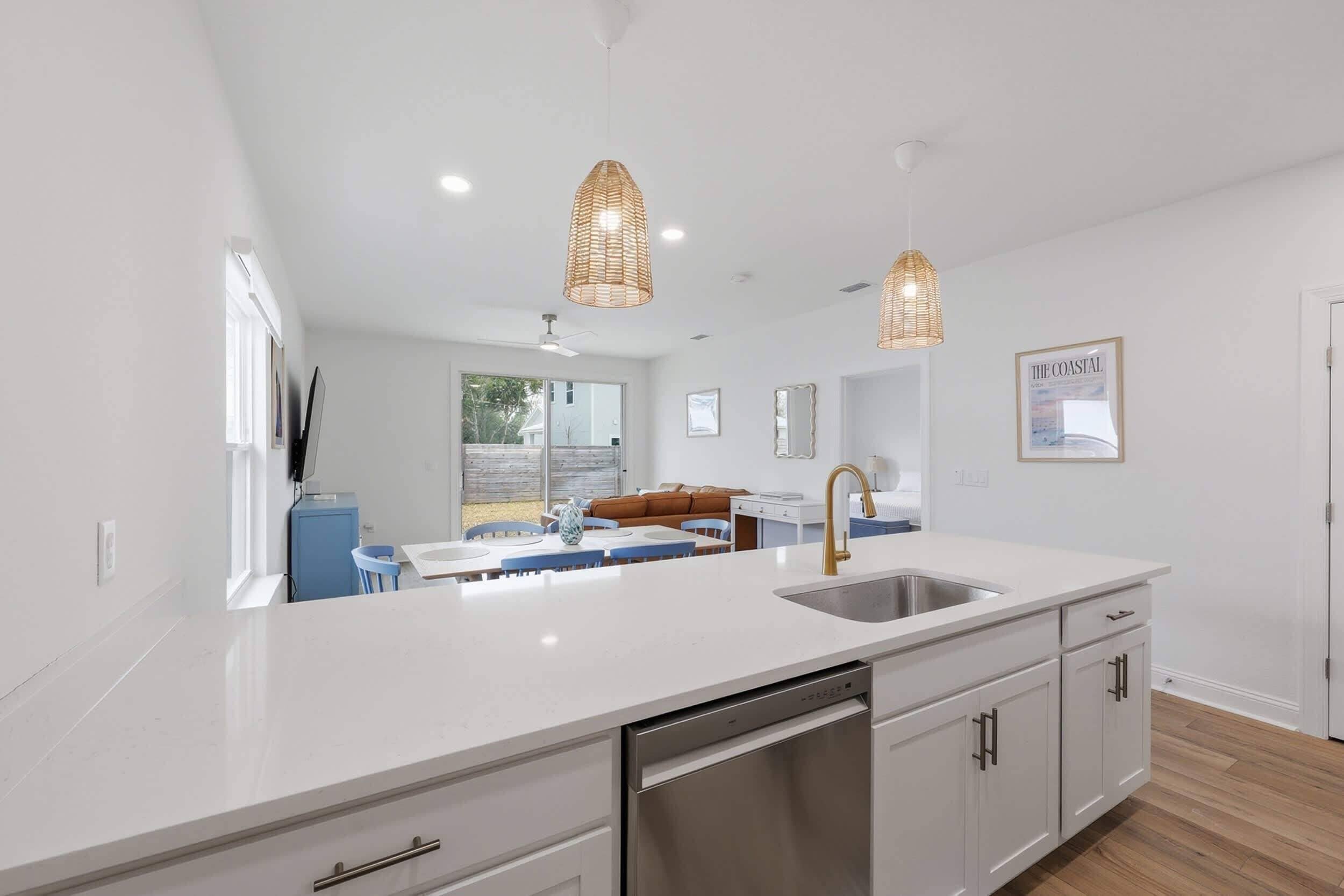Una cocina moderna con muebles blancos, un lavamanos de acero inoxidable y un lavavajillas. Hay dos lámparas colgantes, un ventanal y un cuadro en la pared.