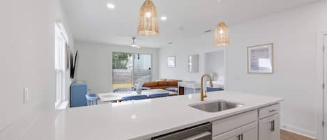 Una cocina moderna con muebles blancos, un lavamanos de acero inoxidable y un lavavajillas. Hay dos lámparas colgantes, un ventanal y un cuadro en la pared.