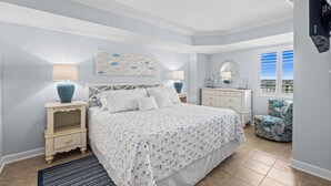 Un dormitorio con cama, mesitas de noche, cómoda, silla, ventana y cuadros en la pared.