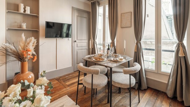 A dining area with a round table, four chairs, a TV on the wall, and a window with curtains. - 1523 Verneuil (Paris)