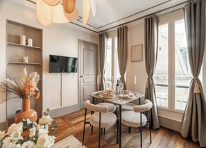 A dining area with a round table, four chairs, a TV on the wall, and a window with curtains. - 1523 Verneuil (Paris)