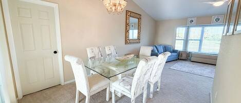 Uma sala de jantar com mesa de vidro e cadeiras brancas, um lustre, um sofá e uma janela com persianas.