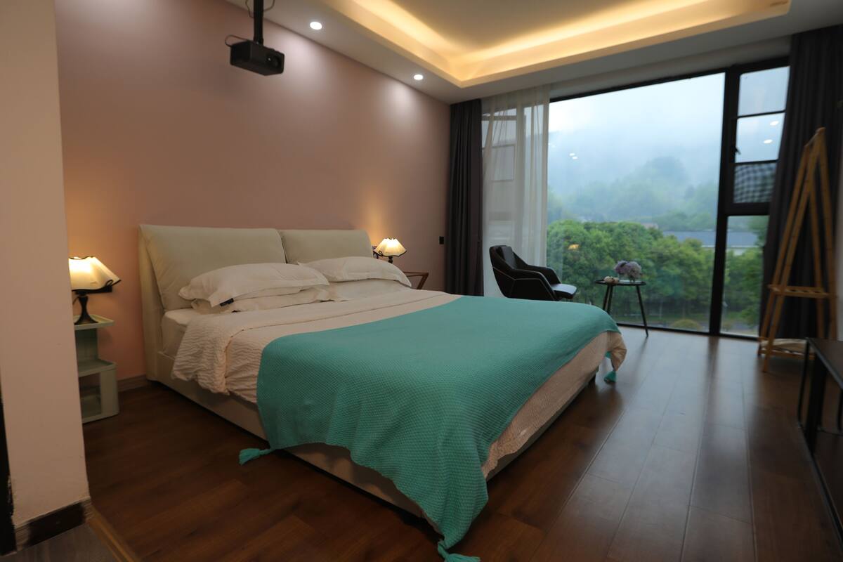 Un dormitorio con cama, lámparas de noche, una silla, una mesita y vistas al exterior.