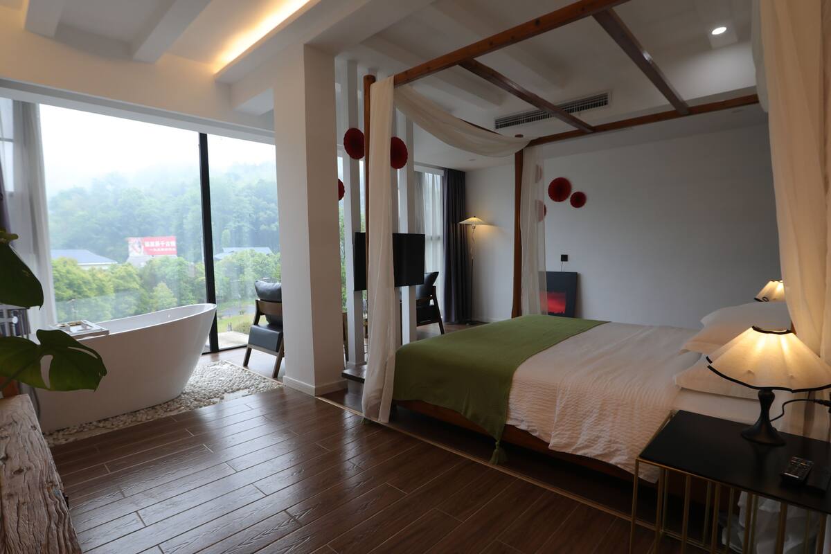 Un dormitorio moderno con un ventanal grande, una bañera, una cama con manta verde y una mesita de noche con lámpara.