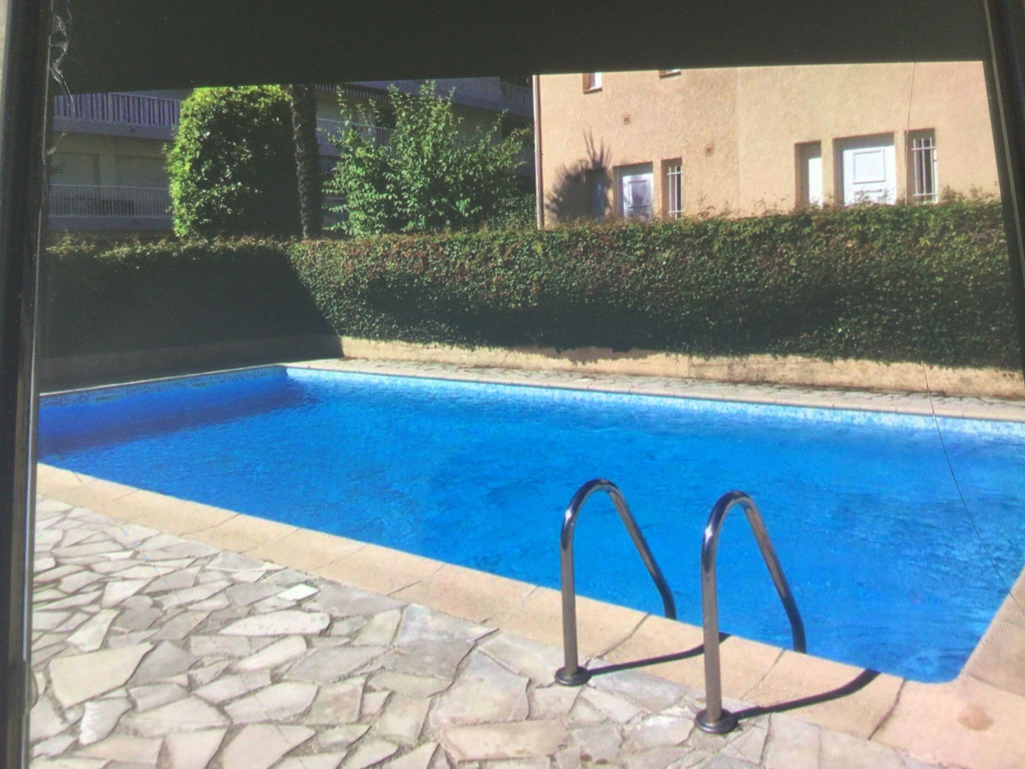 A swimming pool with stone tiles and metal ladders, surrounded by a hedge and a building with windows.