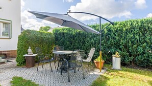 A patio with a table and chairs, a large umbrella, and a hedge in the background.