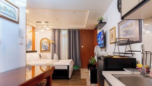 Habitación de hotel con cama, mesita de noche, pequeño escritorio, silla, televisor, ventilador, zona de cocina con lavamanos y piso de madera.