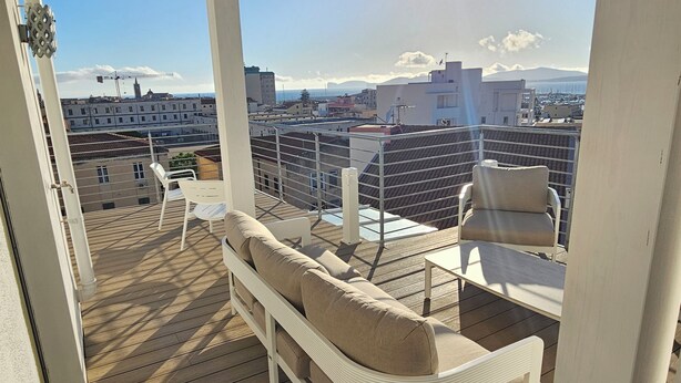 A balcony with a sofa and chairs, overlooking a cityscape with buildings and mountains. - Lecasinedigiò1 Spacious penthouse with large terrace overlooking the sea (Alghero)