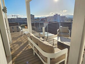 A balcony with a sofa and chairs, overlooking a cityscape with buildings and mountains.