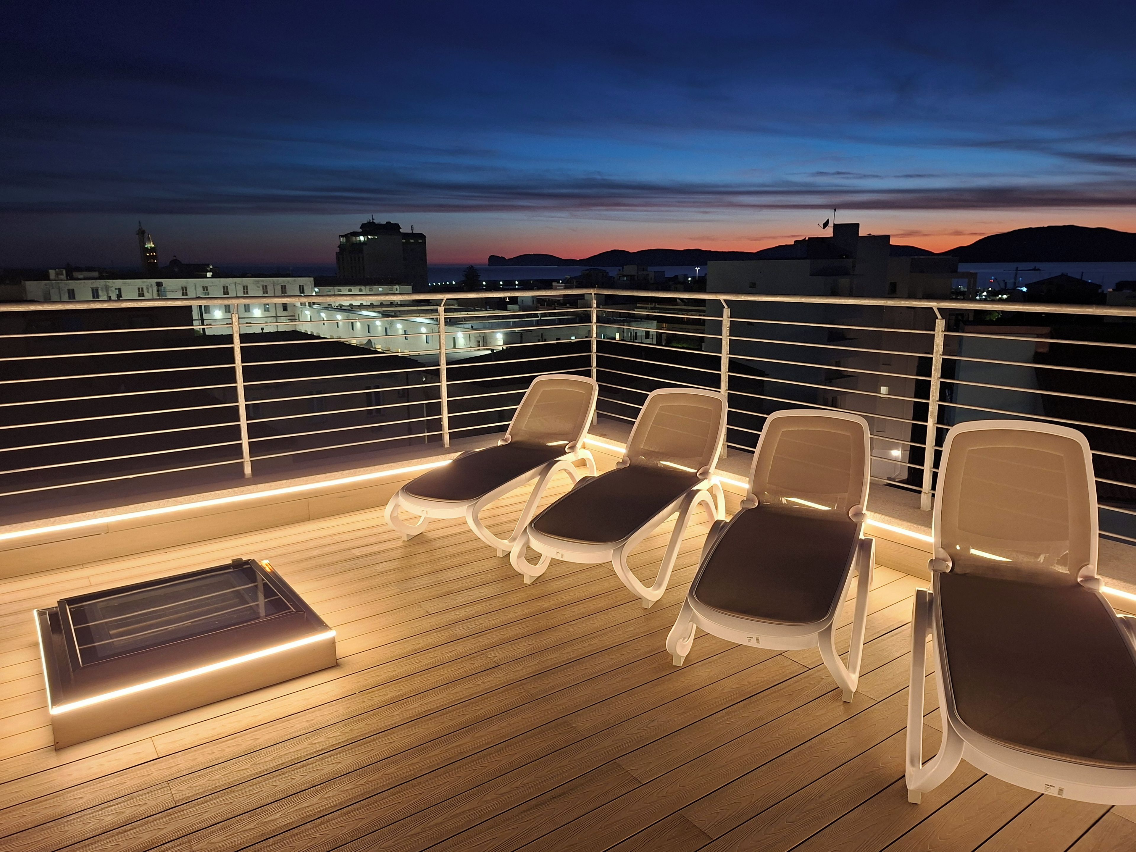 En terrasse med fire liggestole og en solseng, med udsigt over en by ved solnedgang.