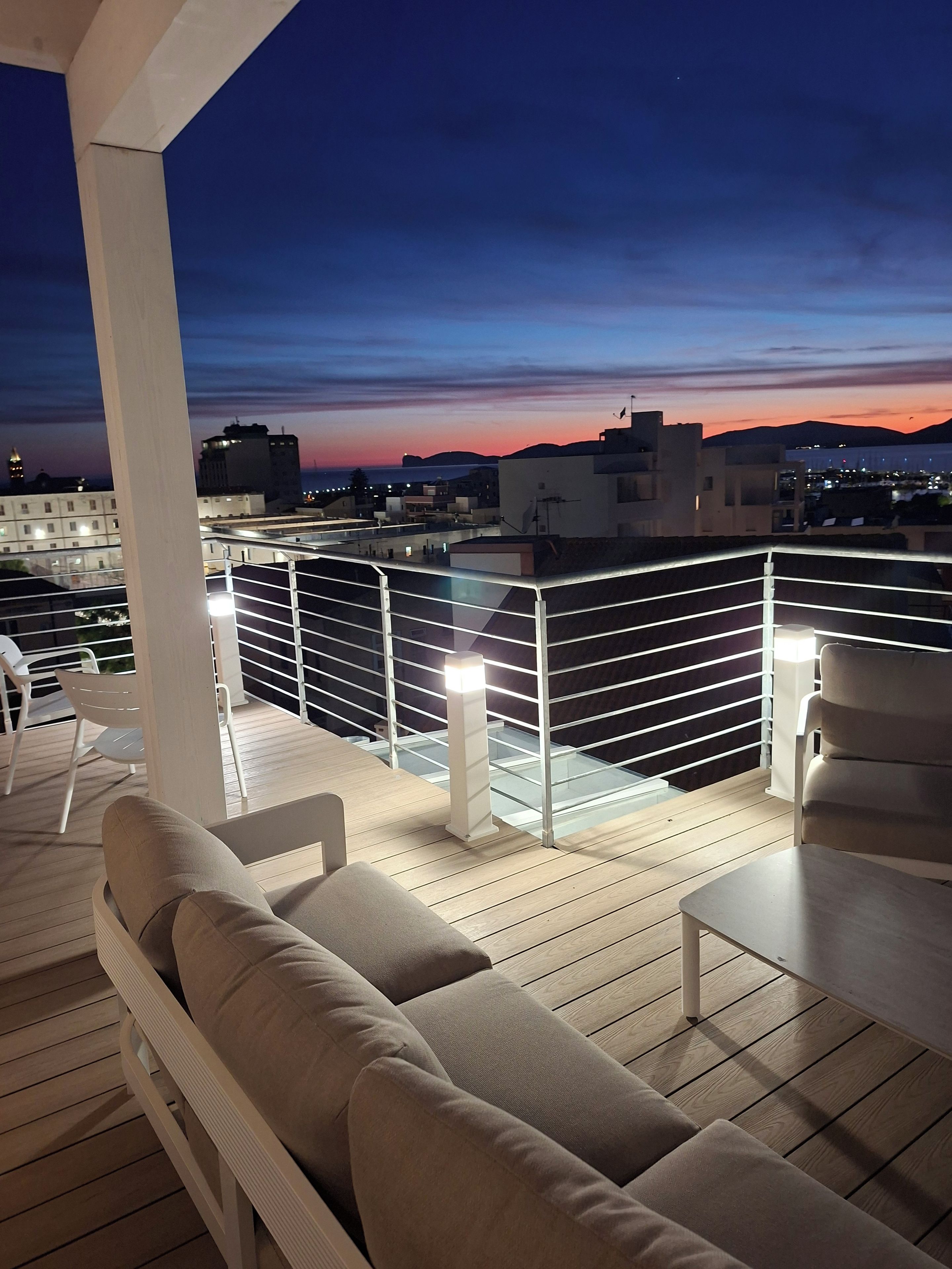 A rooftop terrace with a view of the city skyline at dusk, featuring a railing, seating area, and a table.