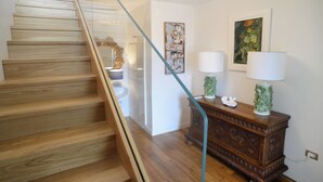 A wooden staircase with a glass railing, a white wall with framed artwork, and a wooden cabinet with two lamps. - Lecasinedigiò1 Spacious penthouse with large terrace overlooking the sea (Alghero)