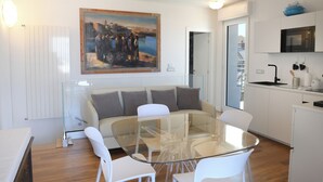 A modern living room with a glass dining table, white furniture, a large painting, and a kitchen area. - Lecasinedigiò1 Spacious penthouse with large terrace overlooking the sea (Alghero)