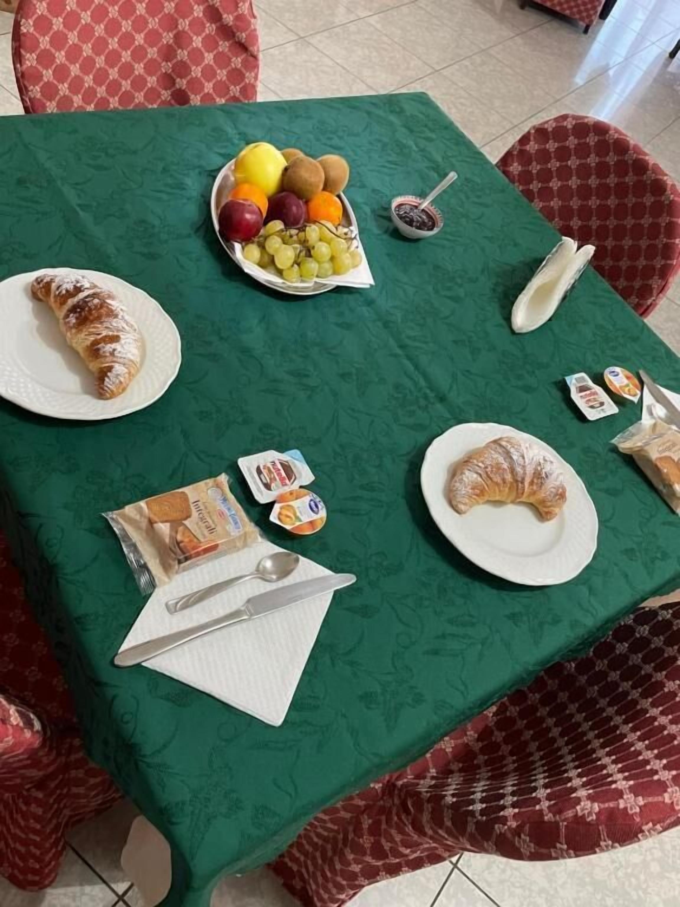 A table set with croissants, fruits, jam, and condiments, with a green tablecloth and red patterned chairs.