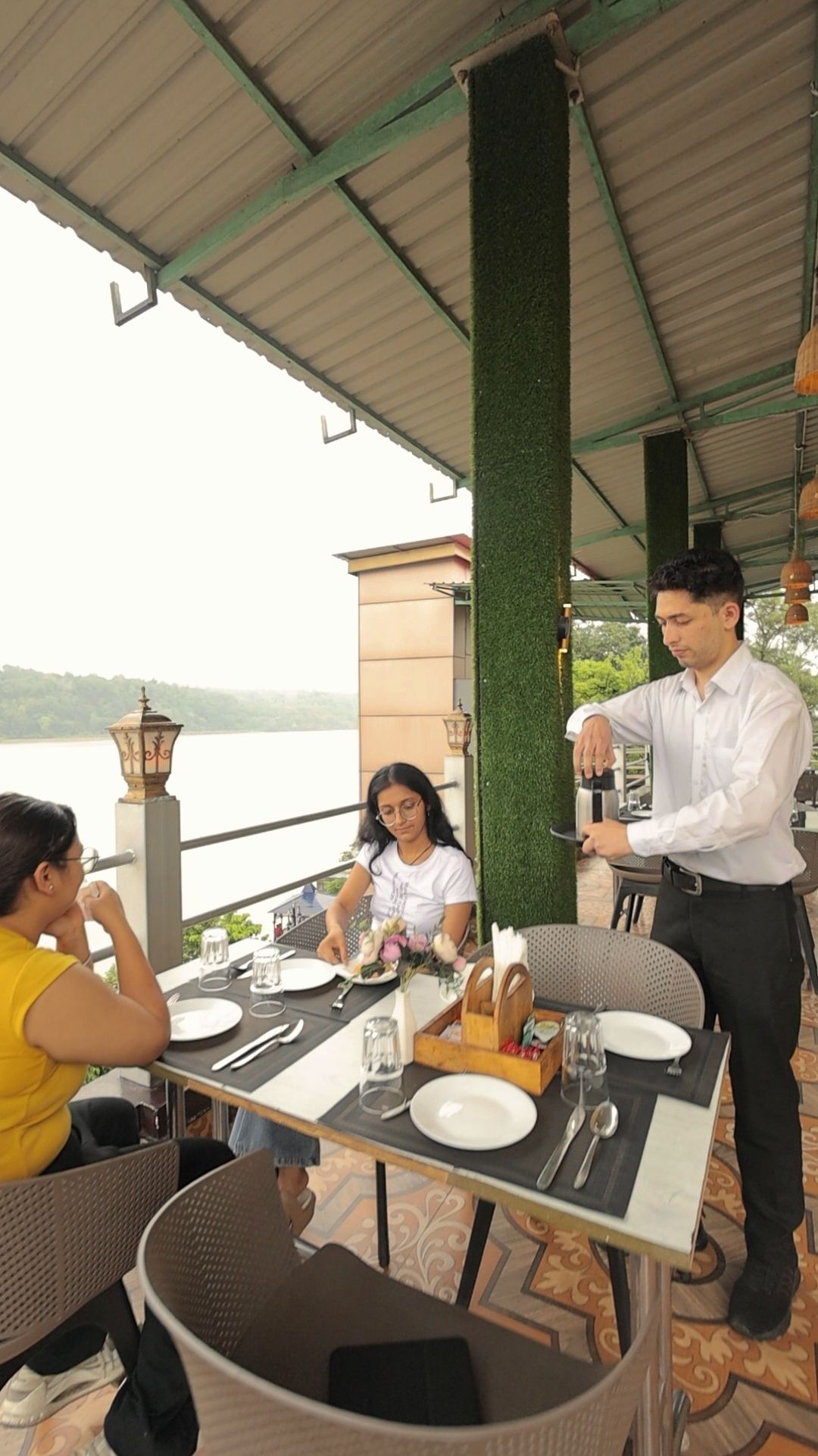 An outdoor dining area with tables and chairs, a waiter attending to guests, and a view of a body of water.