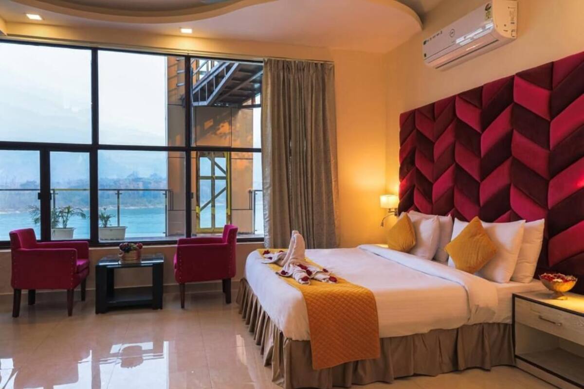 Habitación de hotel con un ventanal grande, una cama con cabecera roja, dos sillas rojas, una mesita y una mesita de noche con una lámpara.
