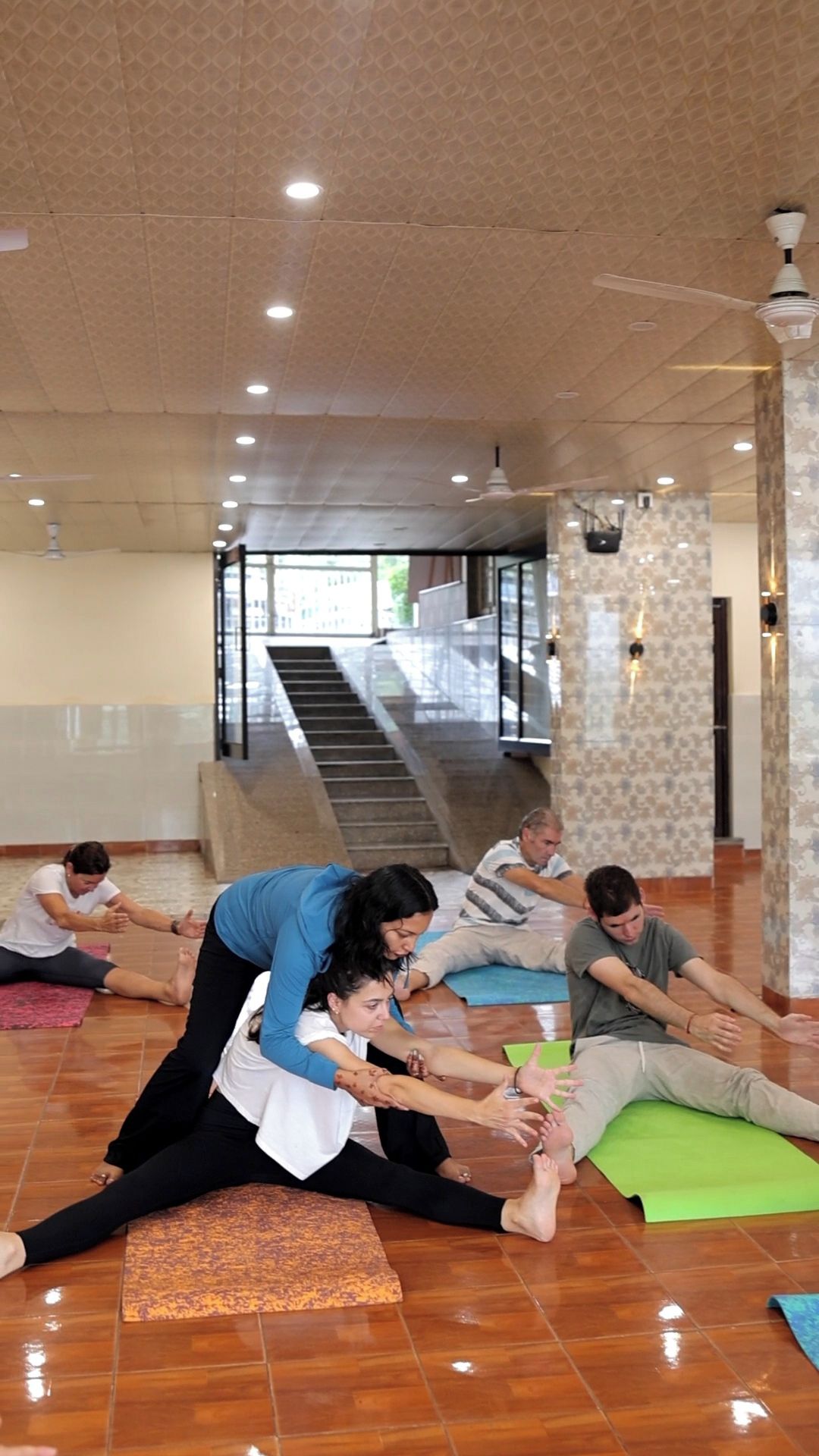 A group of people stretching on mats in a spacious room with a high ceiling and a staircase in the background.