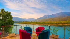A terrace with a view of a river and mountains, furnished with colorful chairs and a small table. - The Shanti Naturecure on Ganga (Rishikesh)