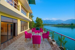 A hotel balcony with a table and chairs, a view of a lake, and mountains in the background.
