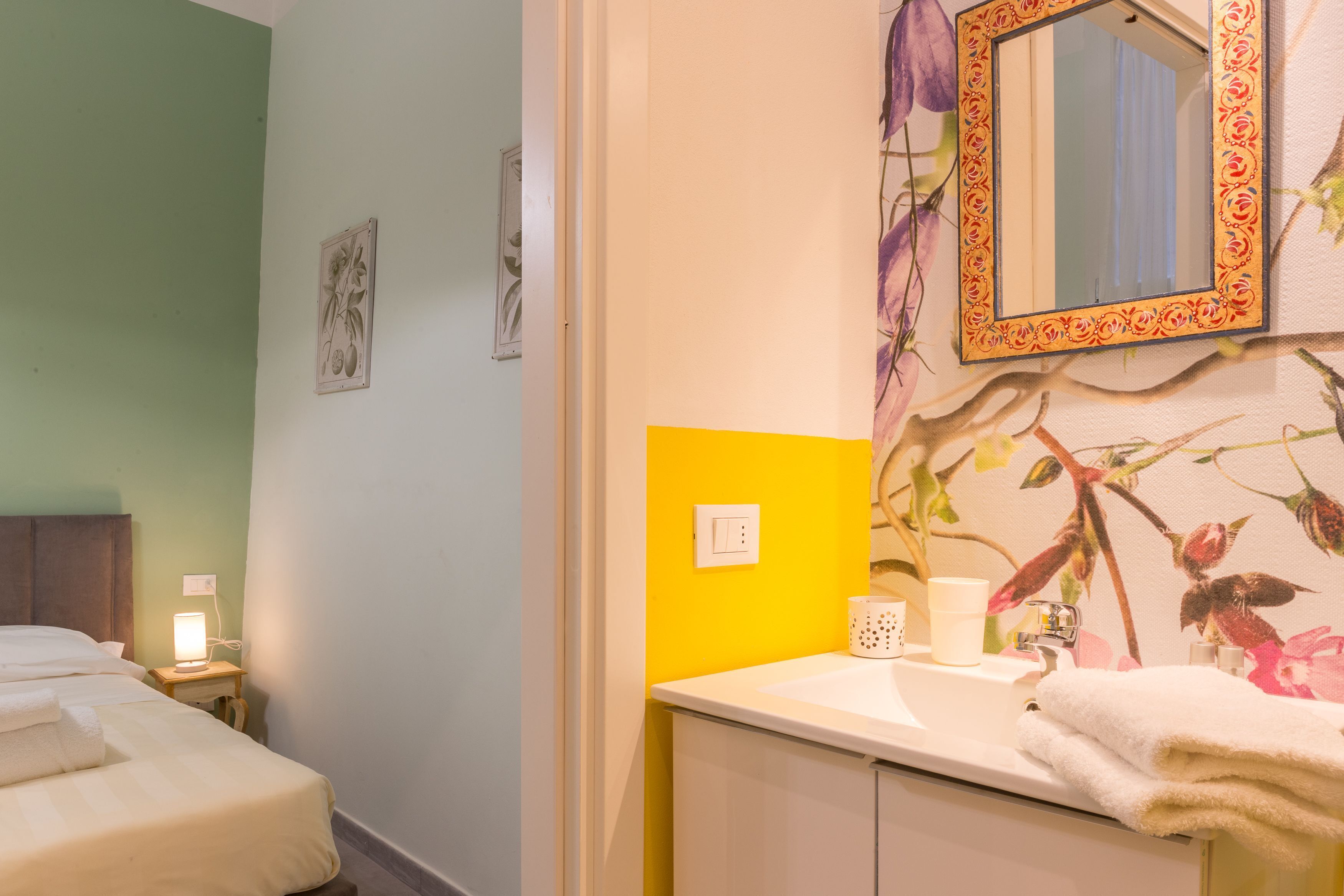 Une chambre d’hôtel avec un lit, une table de chevet avec une lampe, une salle de bain avec un lavabo, un miroir et un papier peint à motifs floraux.