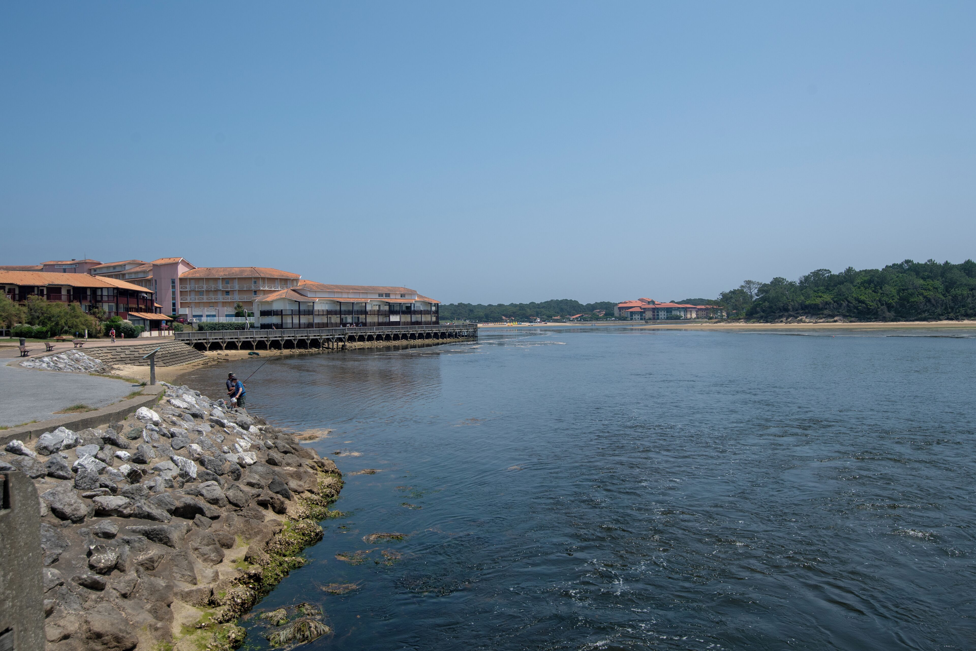 A riverside hotel with a stone pier, a bridge, and buildings with red roofs.
