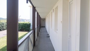 Un pasillo con balcón, paredes blancas y vista a zonas verdes y edificios.