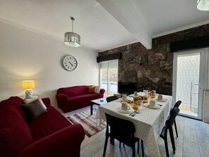 Ein Wohnzimmer mit einem roten Sofa, einem Esstisch gedeckt mit Frühstücksutensilien, einer Uhr an der Wand und einer Steinsäule.