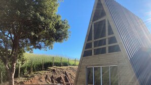 A modern, triangular wooden structure with large windows, a tree, and a grassy area with a fence and a wheelbarrow. - Paraitinga Glamping  (São Luiz do Paraitinga)