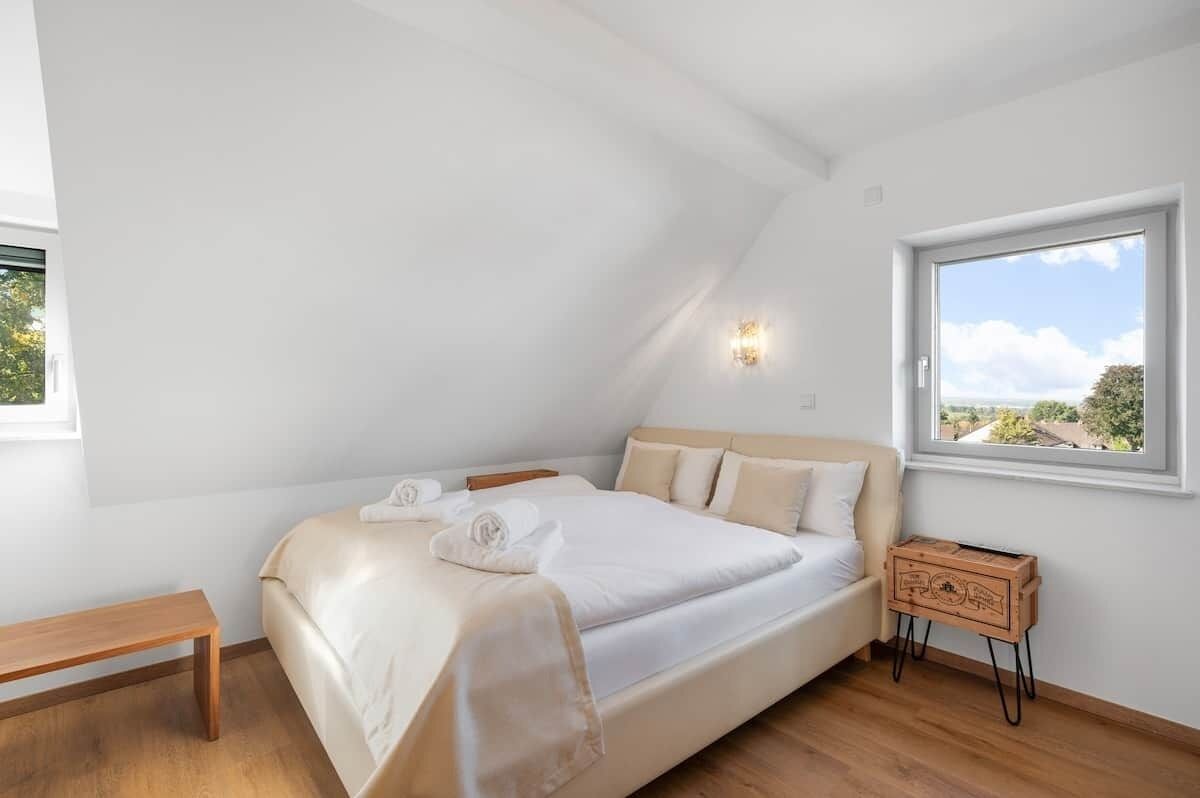 Quarto com cama, um banco, uma mesa de cabeceira, janela e luminária fixada na parede.