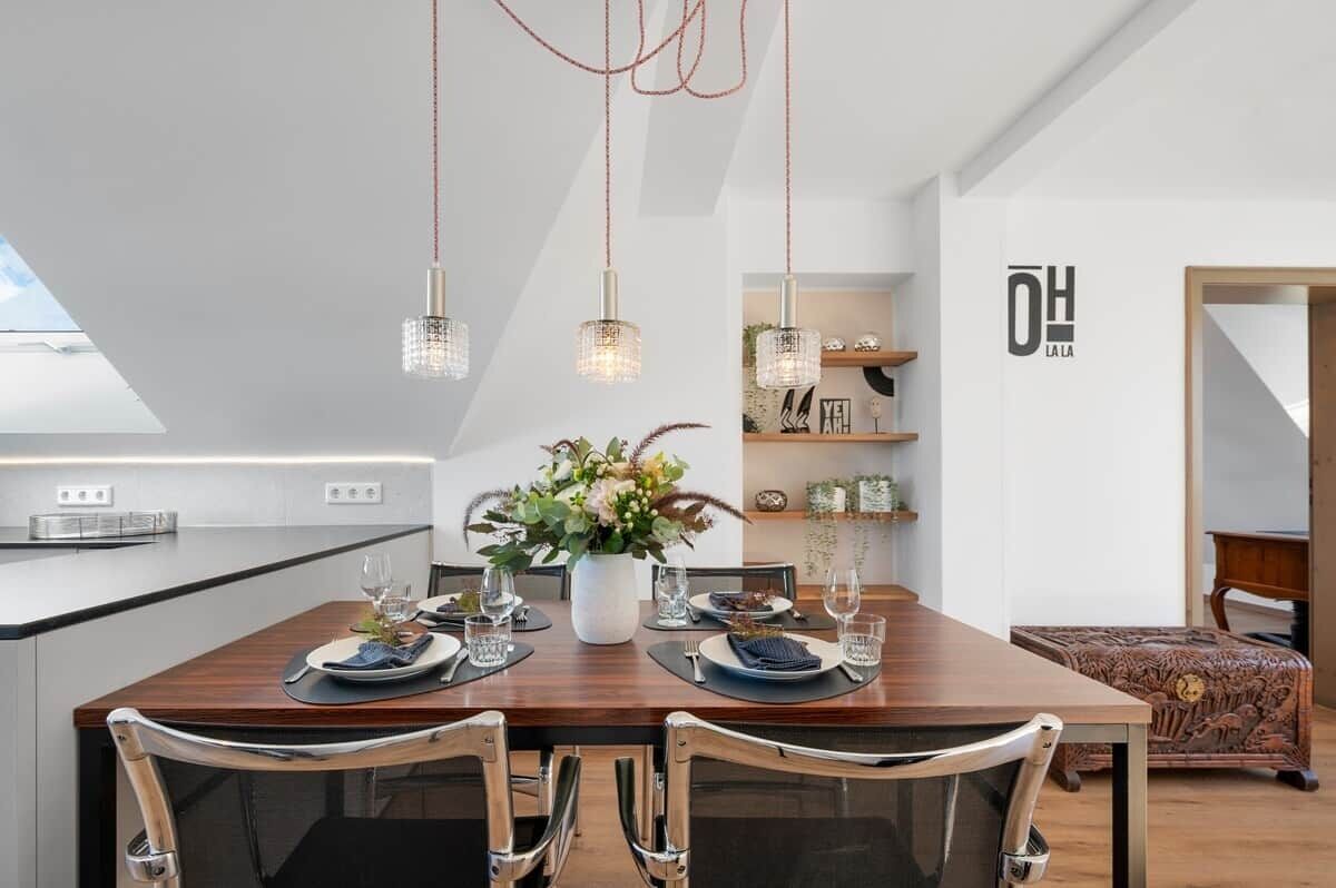 A modern kitchen with a dining area, pendant lights, a vase with flowers, and a sign on the wall.