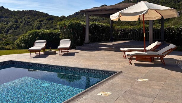 Una piscina con sillas de descanso y sombrillas, rodeada por un jardín ajardinado y con montañas al fondo.