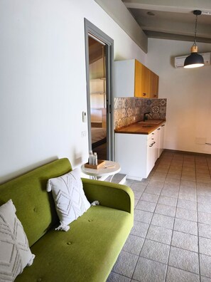 Un espai habitable reduït amb un sofà verd, una taula rodona petita, una zona de cuina amb salabost i una lluminària penjant del sostre.