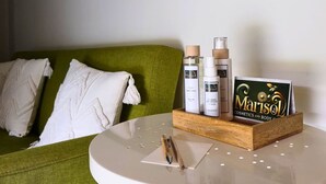 Un sofá verde con almohadillas blancas, una mesa blanca con una bandeja que contiene botellas y una tarjeta, y una pared blanca.