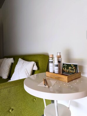 A green sofa with white pillows, a white table with a tray holding bottles and a card, and a white wall.