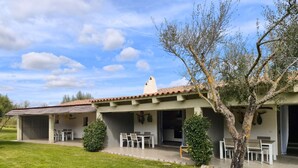 Un edificio de dos pisos con un patio, mobiliario exterior y un árbol en primer plano.