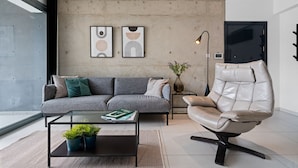 Un salon moderne avec un canapé gris, un fauteuil en cuir, une table basse en verre agrémentée de plantes et deux œuvres d’art abstraites accrochées au mur.