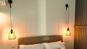 Een net opgemaakt bed met witte lakens en bruine accenten, twee hanglampen, een klein rond tafeltje en een schakelaar op de muur.