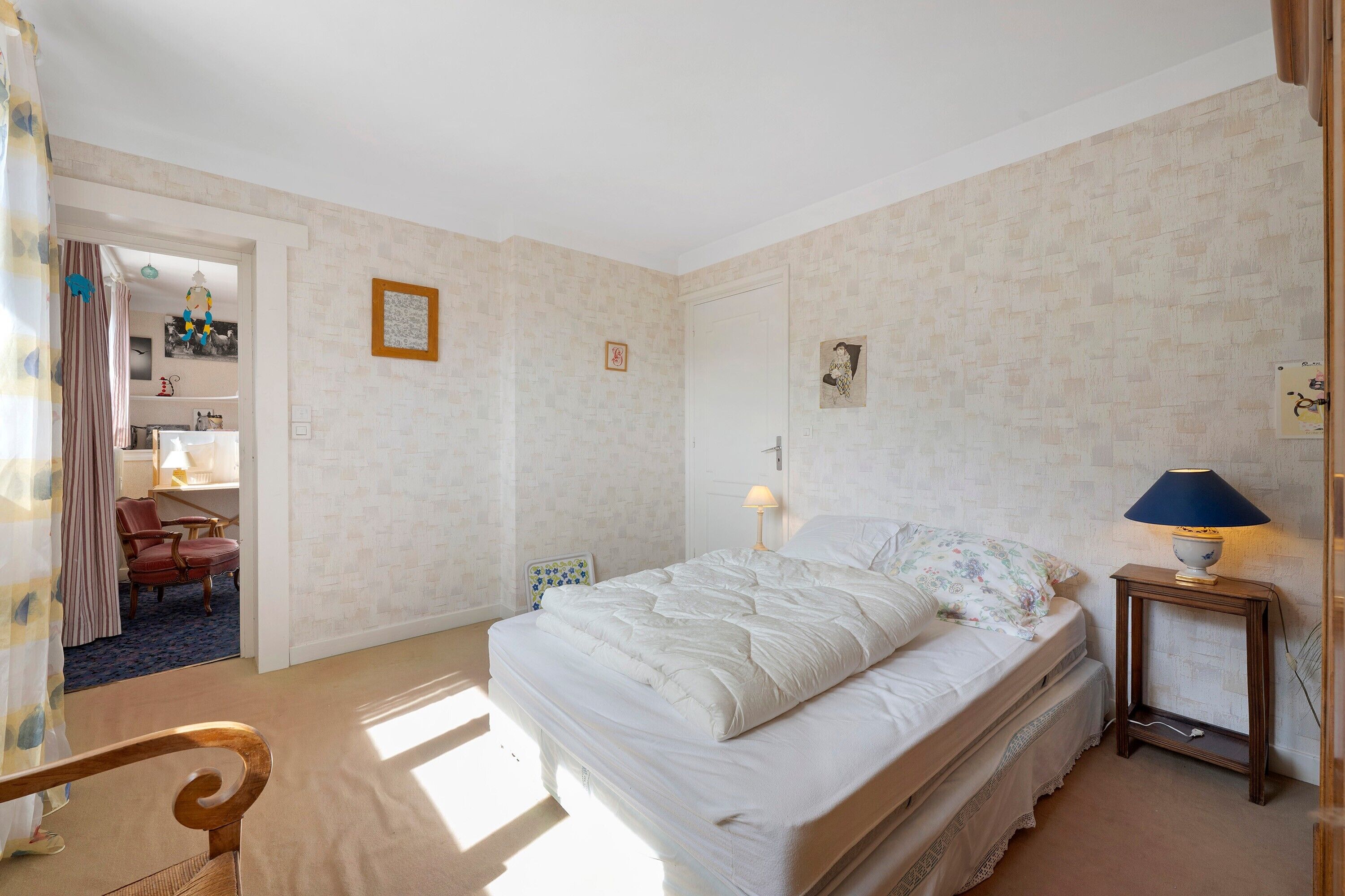 Un dormitori amb un llit, una nit, una làmpada, una cadira i una porta que condueix a una altra habitació.