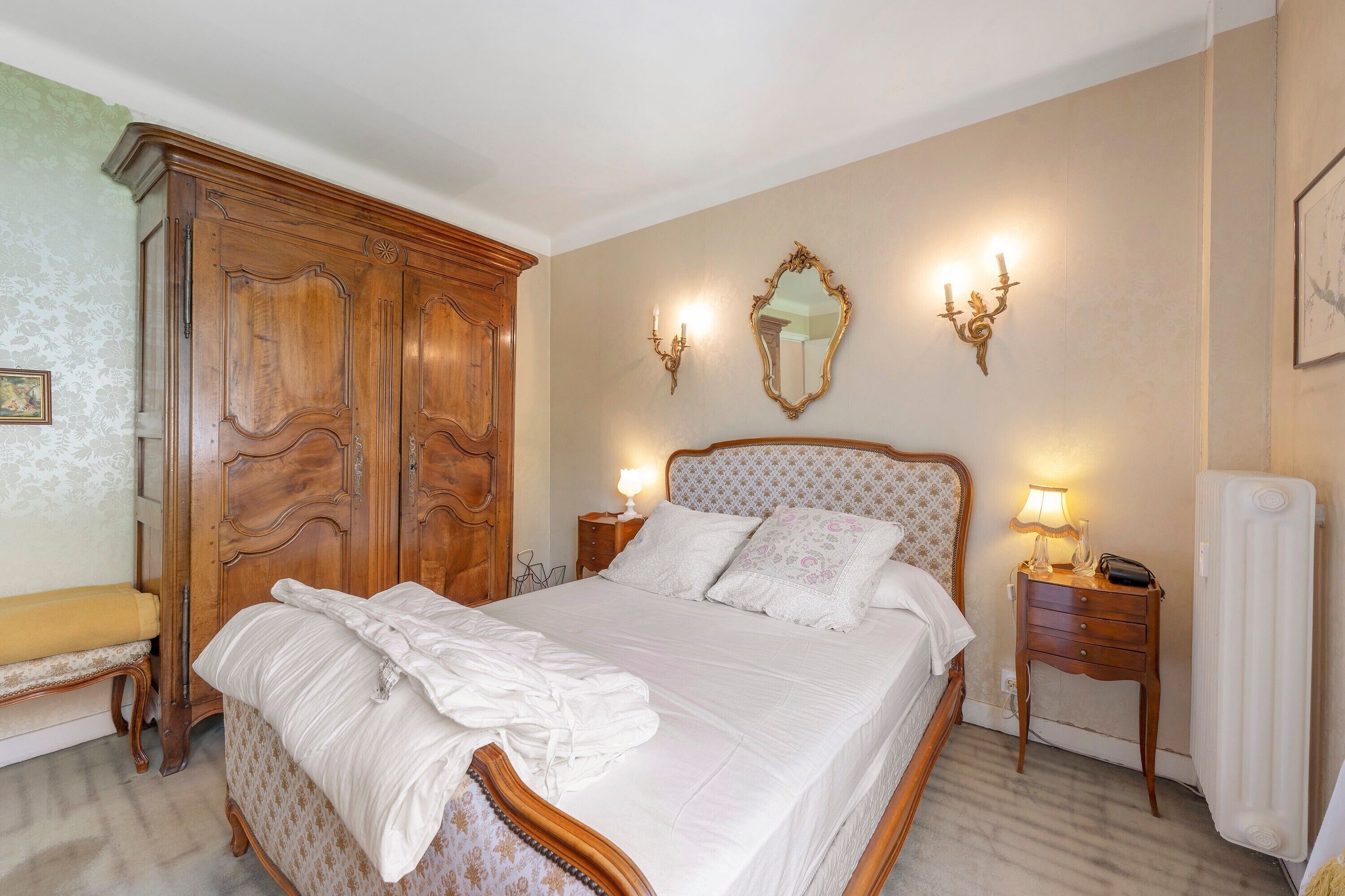Una camera da letto con un'ampia armadio in legno, un letto con lenzuola bianche, due lampade da comodino, uno specchio e un termosifone.