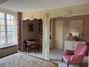 Une chambre à coucher avec un lit, un fauteuil rose, une chaise en bois, une table recouverte d’une nappe verte, une bibliothèque et une fenêtre avec des rideaux.