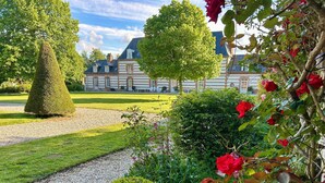 Un jardin bien entretenu, avec un chemin de gravier, des haies soigneusement taillées et, au fond, un grand bâtiment au style architectural traditionnel.