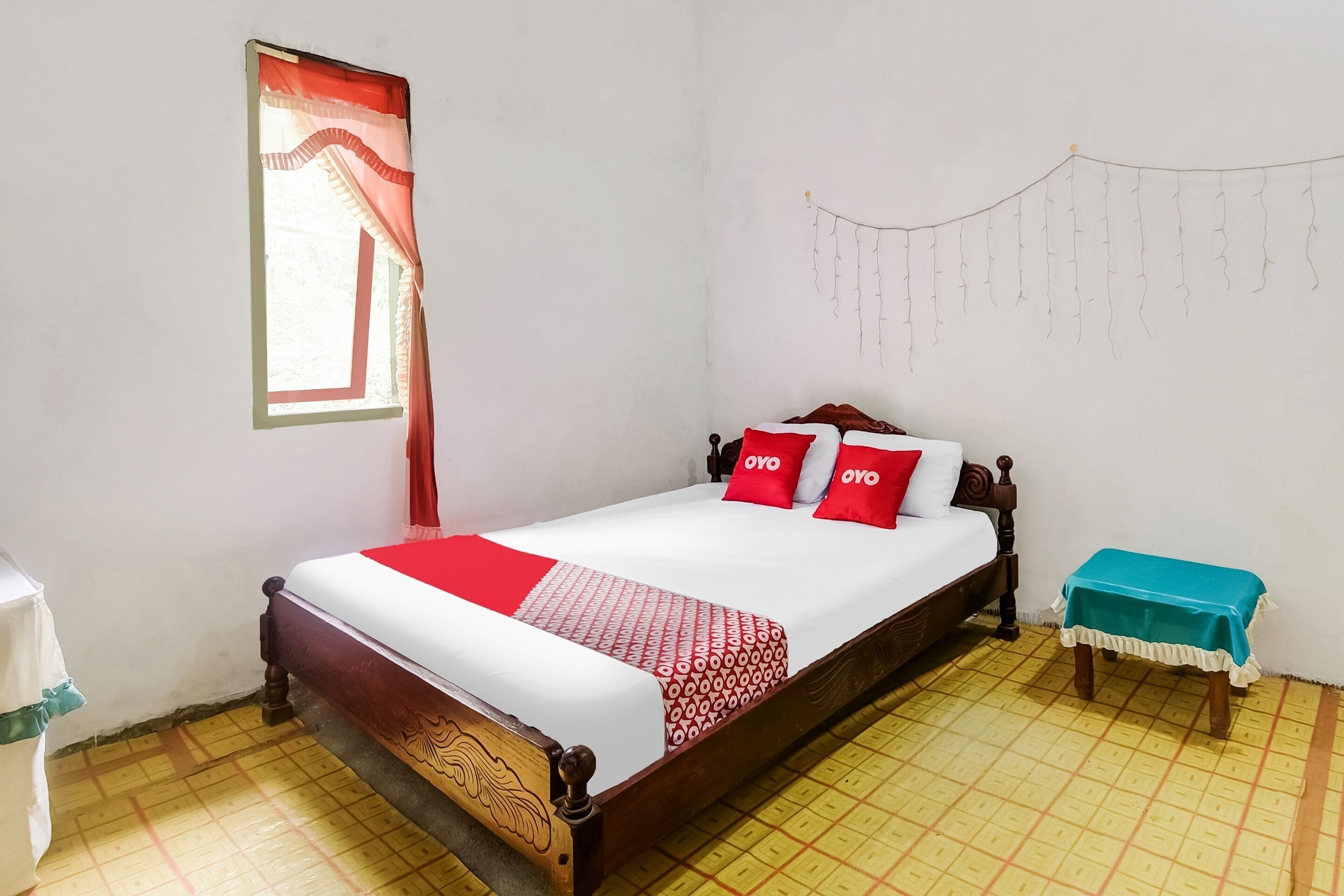 A single bed with a wooden frame, a red and white bedspread, two red pillows, a small bedside table with a teal cloth, and a window with red curtains.