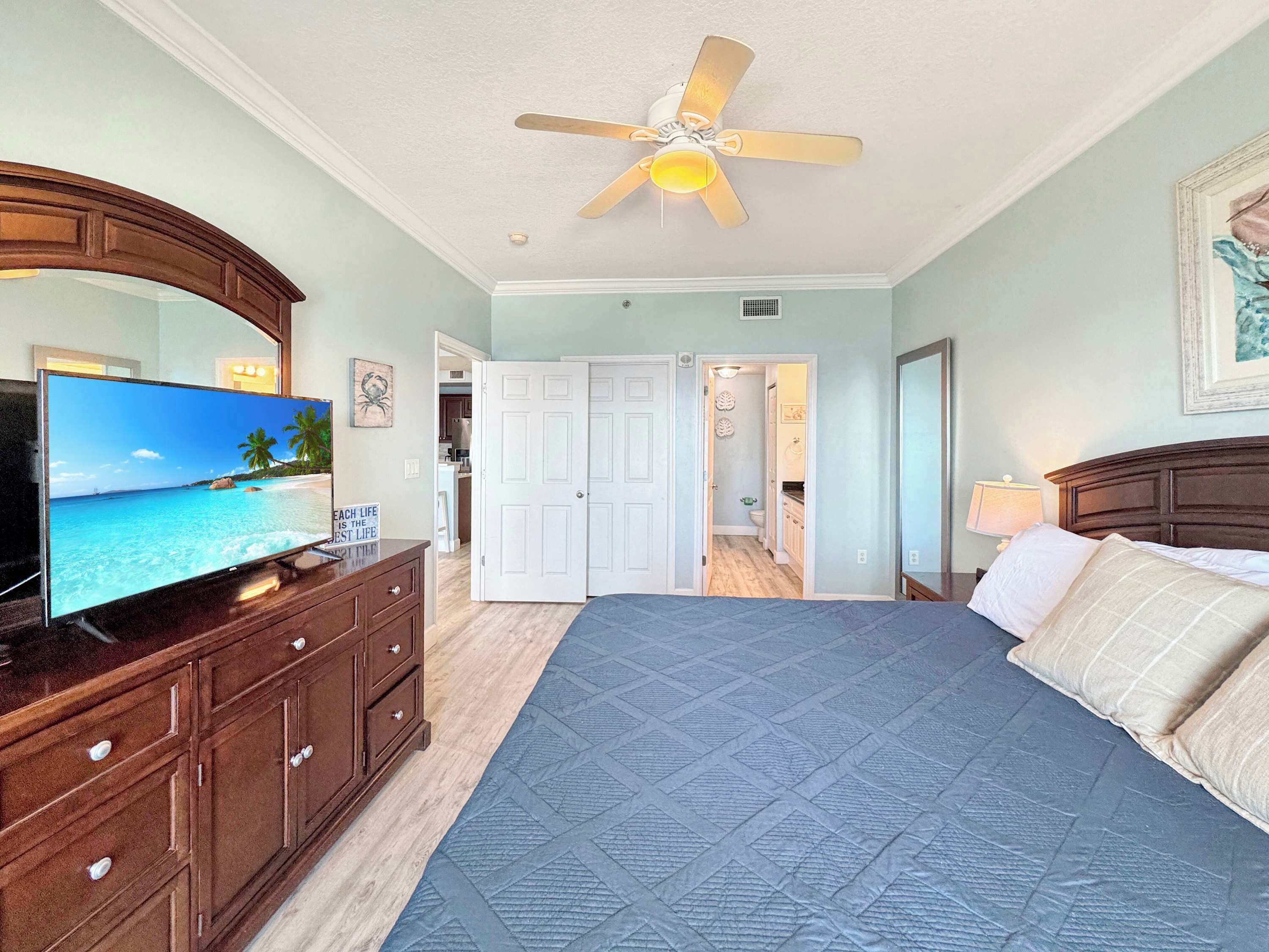 A bedroom with a bed, a television, a ceiling fan, a dresser, and a door leading to a bathroom.