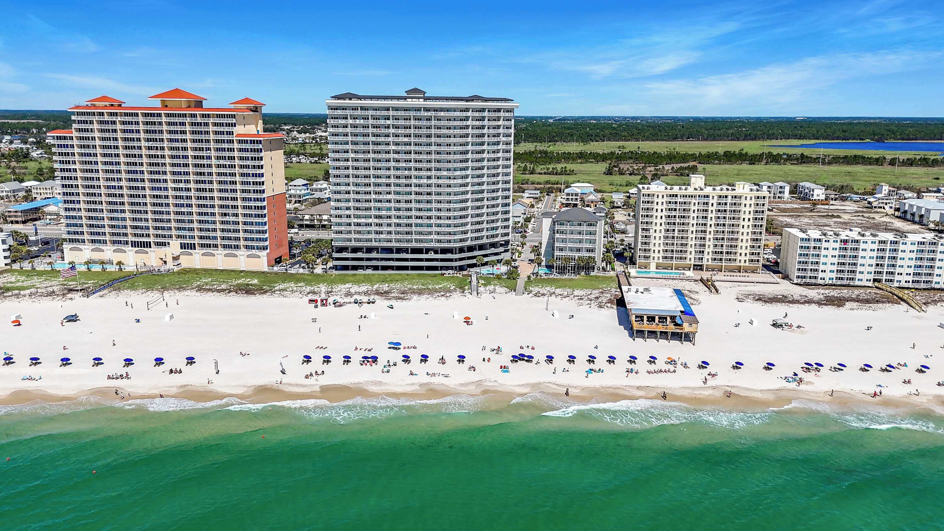 A beachfront hotel with multiple high-rise buildings, a sandy beach with umbrellas, and a clear view of the ocean.