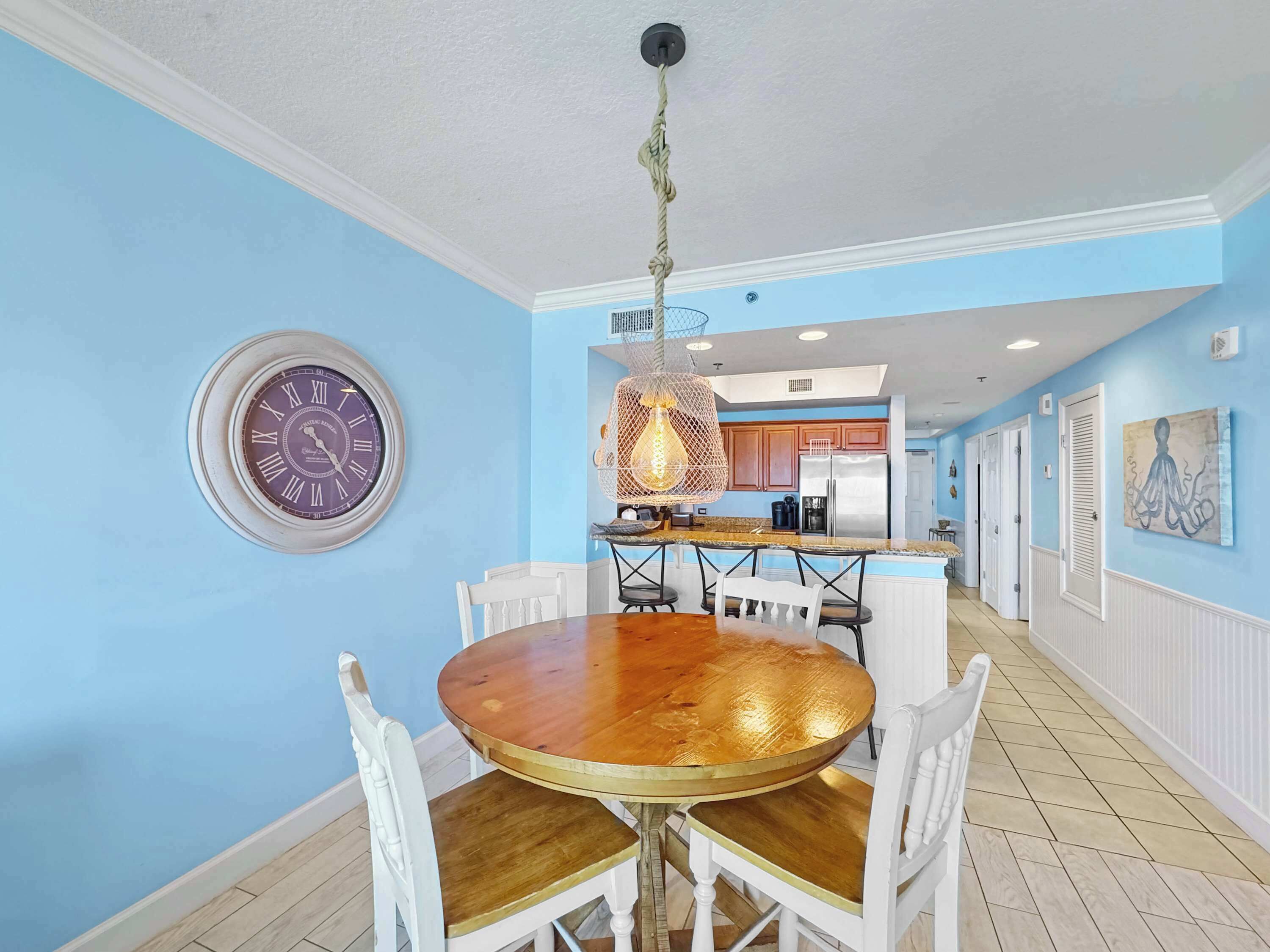 A dining area with a round wooden table, white chairs, a wall clock, and a kitchen in the background.