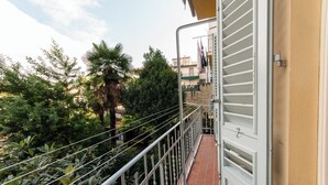 En balkon med gelænder i metal, flisegulv og udsigt til grønne områder og bygninger i baggrunden.