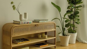 Een rattenboekenplank met boeken en een plant, tegen een effen muur en houten vloer.